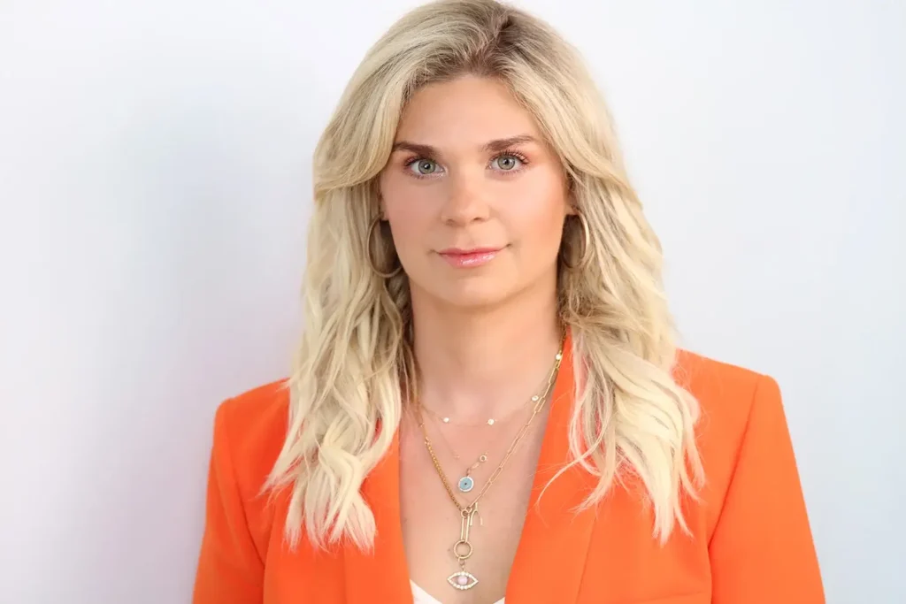 Professional Headshots NYC: A blonde woman in a vibrant orange blazer and gold jewelry against a white background.