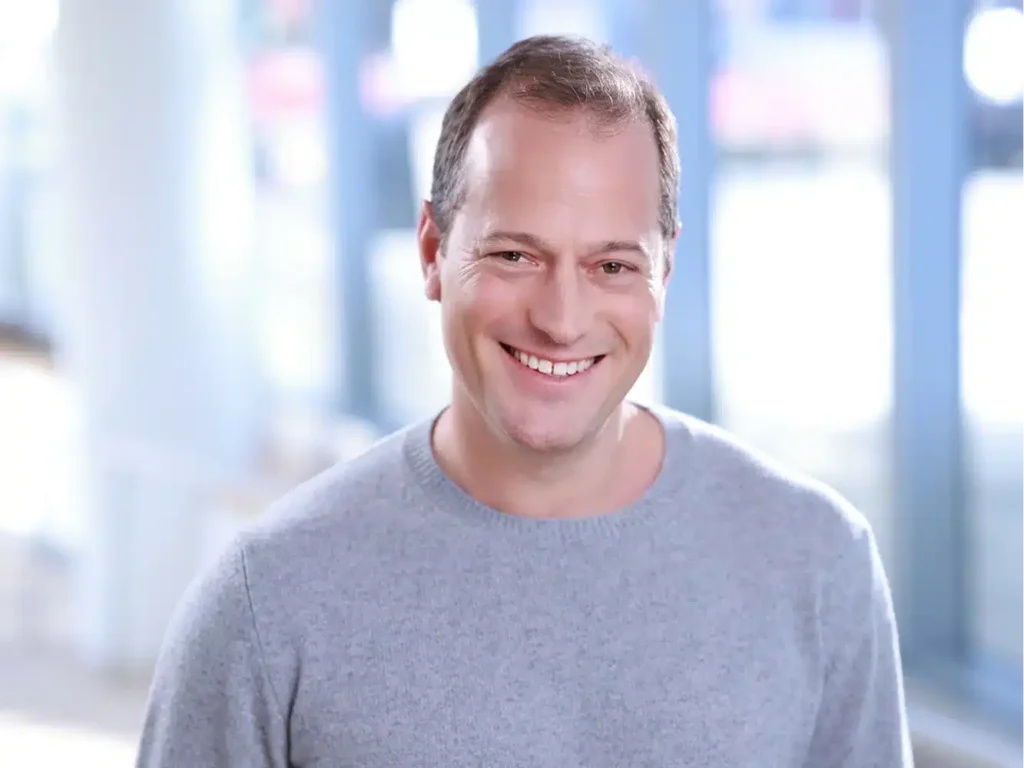 Corporate headshots in NYC: a smiling man in a grey sweater against a brightly lit office background, casual yet professional.