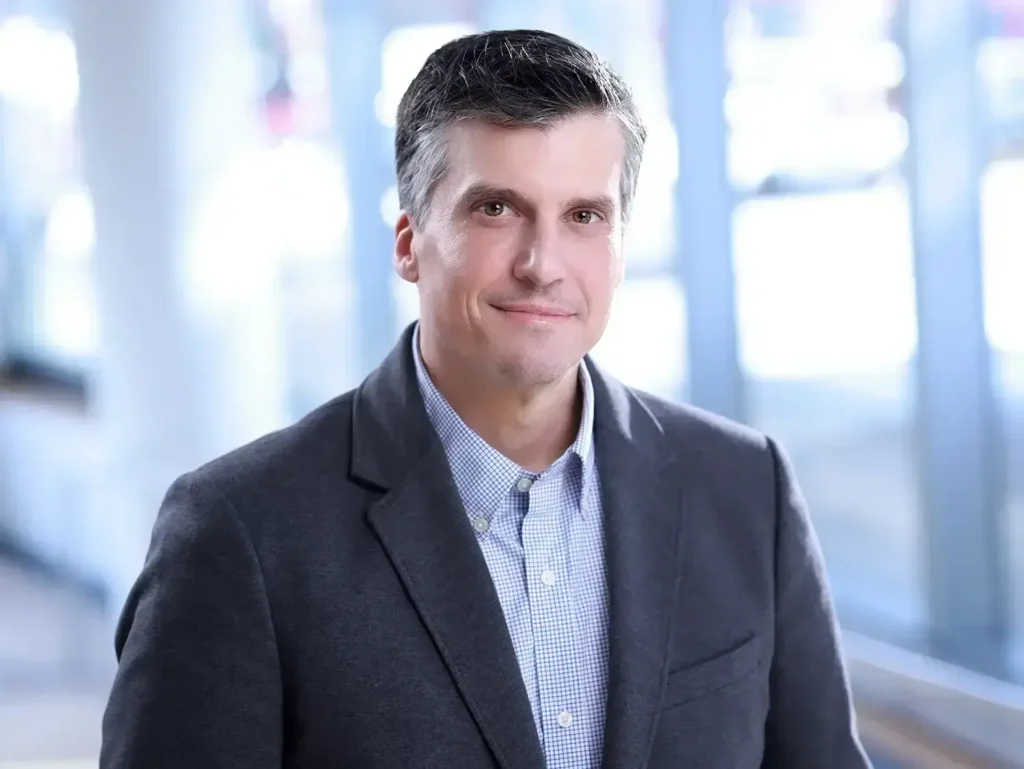 Experienced male executive smiling for his corporate headshots NYC with a blurred city office background.