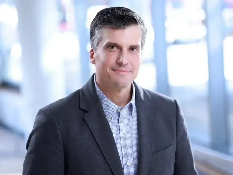 Experienced male executive smiling for his corporate headshots NYC with a blurred city office background.